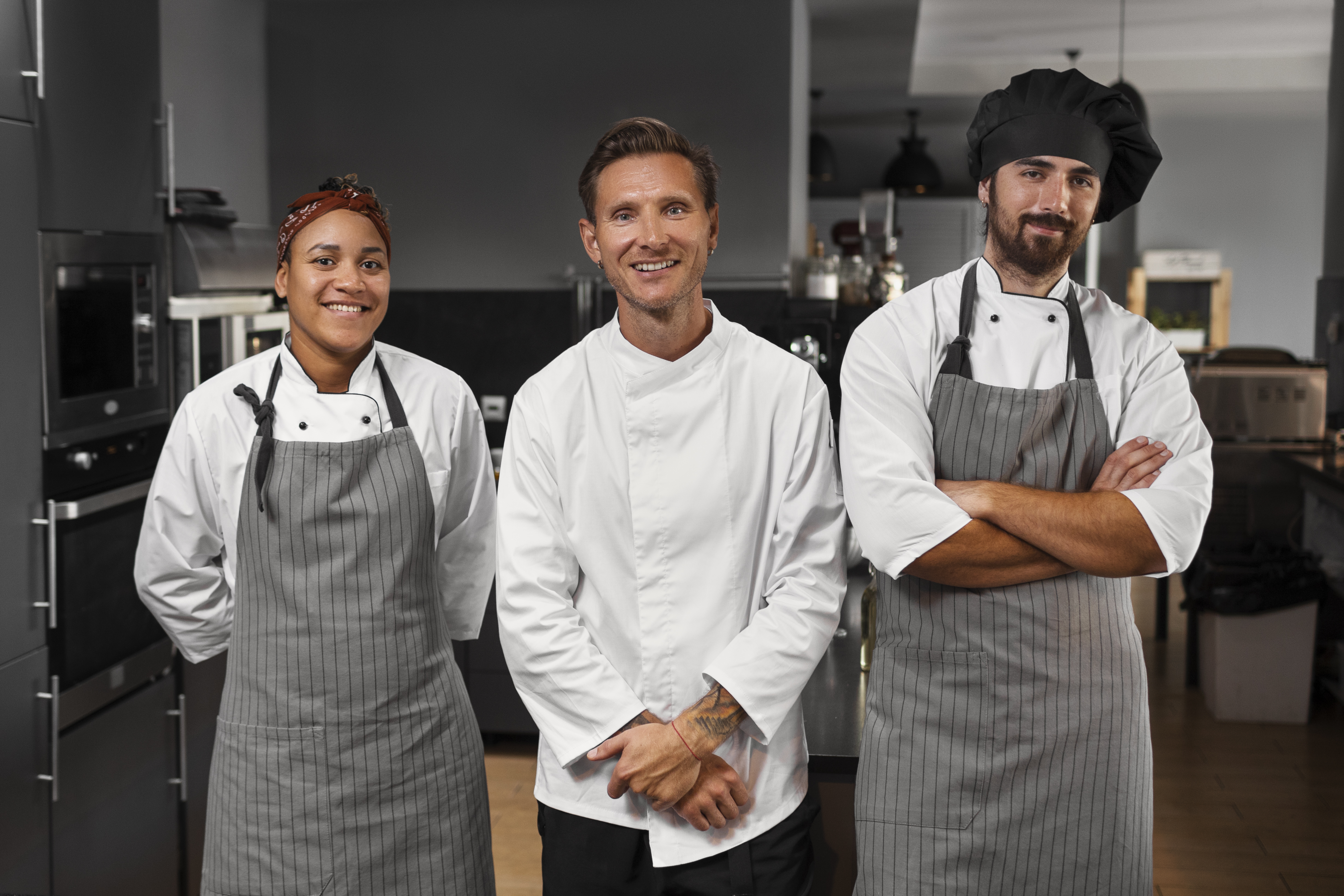 Três chefs de cozinha posando em uma cozinha profissional, todos usando uniforme branco e avental cinza.